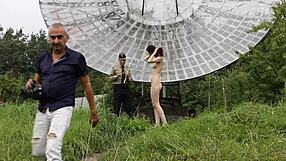 In a public outdoor setting near an old space radar, a brunette with natural tits poses nude