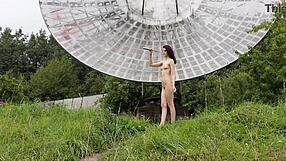 In a public outdoor setting near an old space radar, a brunette with natural tits poses nude