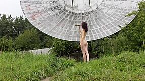 In a public outdoor setting near an old space radar, a brunette with natural tits poses nude